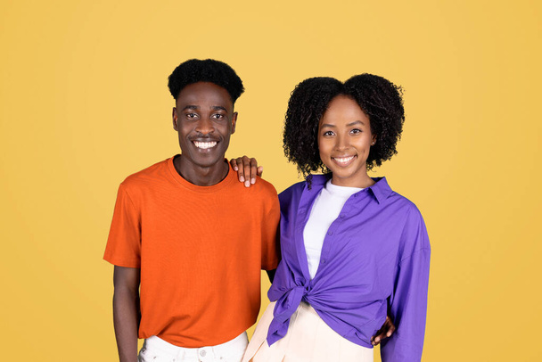 Una joven sonriente con una blusa púrpura y un joven con una camisa naranja de pie hombro a hombro, mirando a la cámara con un comportamiento feliz sobre un fondo amarillo - Foto, Imagen