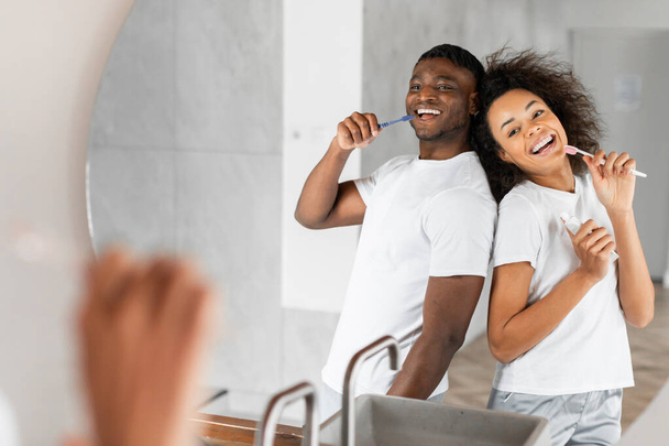 Χαρούμενο παντρεμένο ζευγάρι Αφροαμερικανών με casual ρούχα τραγουδώντας ανέμελα ενώ βουρτσίζουν τα δόντια τους, διασκεδάζοντας μπροστά στον καθρέφτη του μπάνιου, μοιράζοντας στιγμές πρωινής ευτυχίας. Αντιγραφή χώρου - Φωτογραφία, εικόνα