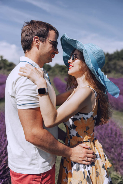 Een gelukkig jong stel loopt door een lavendelweide. Een man en een roodharige vrouw knuffelen elkaar terwijl ze naar elkaar kijken. Een lachende vrouw met een hoed op. Verliefd stel, man en vrouw op het lavendelveld - Foto, afbeelding
