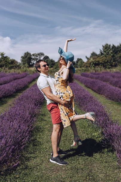 Rakastunut pariskunta, mies ja nainen, kävelevät laventeliniityllä. Mies pitää naista sylissään. Hattupäinen nainen hymyilee ja nostaa kätensä taivasta kohti. Kesäkävely laventelipellon läpi - Valokuva, kuva