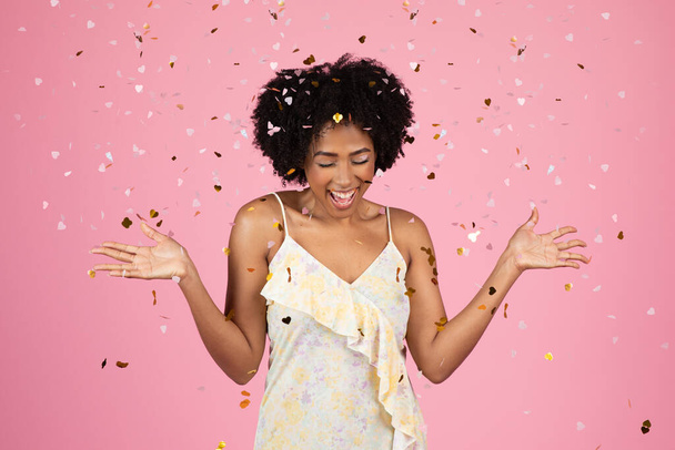 Mujer afroamericana milenaria animada con el pelo rizado riéndose y levantando las manos en el aire, con confeti cayendo a su alrededor durante un momento festivo sobre un fondo rosa - Foto, Imagen