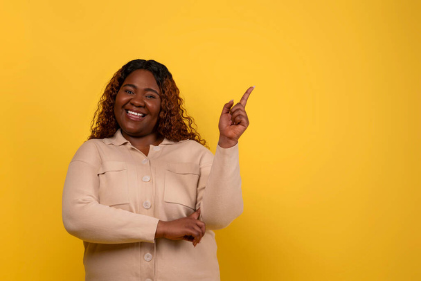 Radosna nadwaga czarna dama wskazująca na puste miejsce na reklamę odizolowaną na żółtym tle studia. Afrykańska pulchna kobieta poleca świetną ofertę, ofertę - Zdjęcie, obraz