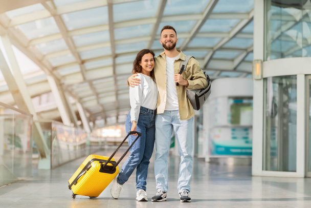 Έτοιμος για ταξίδι. Ευτυχισμένο νεαρό ζευγάρι που ποζάρει στον τερματικό σταθμό του αεροδρομίου, χαρούμενος Χιλιετής άντρας και γυναίκα που στέκεται με βαλίτσα και χαμογελώντας στην κάμερα, περιμένοντας την πτήση, απολαμβάνοντας το ταξίδι μαζί - Φωτογραφία, εικόνα