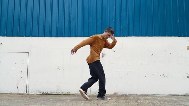 Latynos wyciąga ręce i tańczy przed murem. Motion shot stylowej tancerki lub choreografa w stroju casual praktykujących taniec w stylu hip hop. Sport na świeżym powietrzu 2024. Endeavor. - Zdjęcie, obraz