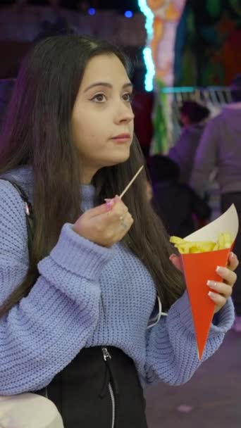 Two girls with serious faces eating potato fries and waffles from food truck in Valencia funfair at night when people passing by in masks - FullHD Vertical video - Footage, Video