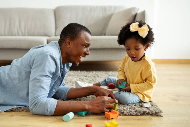Μια όμορφη μαύρη κοπέλα που διασκεδάζει με τον μπαμπά της στο σπίτι. Αξιολάτρευτος Αφροαμερικάνος πατέρας και κόρη κάθονται στο πάτωμα, παίζοντας πολύχρωμα ξύλινα μπλοκ - Φωτογραφία, εικόνα