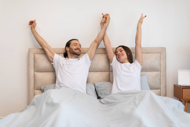 Свежепроснувшаяся пара в белых футболках с поднятыми руками встречает новый день с радостью и энергией в солнечном интерьере спальни - Фото, изображение