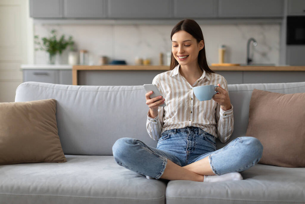 Hymyilevä nuori nainen nauttii rauhallisesta hetkestä kuppi kahvia ja älypuhelinta kädessä, istuu poikkijalkainen mukavalla sohvalla kotona, kopioi tilaa - Valokuva, kuva