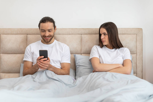 Ο άνθρωπος είναι χαρούμενος που ασχολούνται με το smartphone του, ενώ η γυναίκα δίπλα του κοιτάζει με ανησυχία και δυσαρέσκεια έκφραση, υπαινίσσεται ψηφιακή απόσπαση της προσοχής - Φωτογραφία, εικόνα