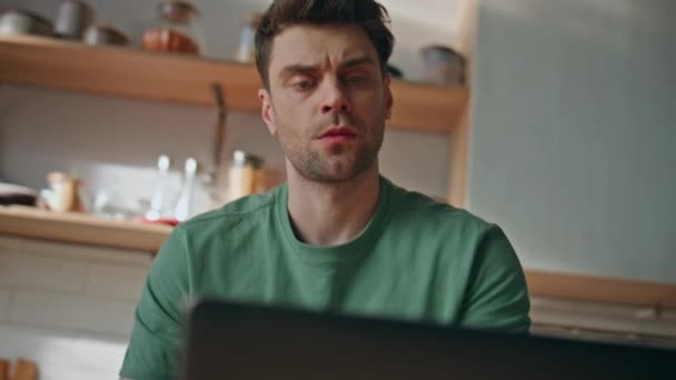 Ciddi bir adam, mutfaktaki bilgisayarla yakından ilgilenir. Tıraşsız adam hafif evde bilgisayar ekranının klavye yazmasını izliyor. Kaşlarını çatan serbest çalışan kahvaltıda e-posta atarak soruları cevaplıyor - Video, Çekim
