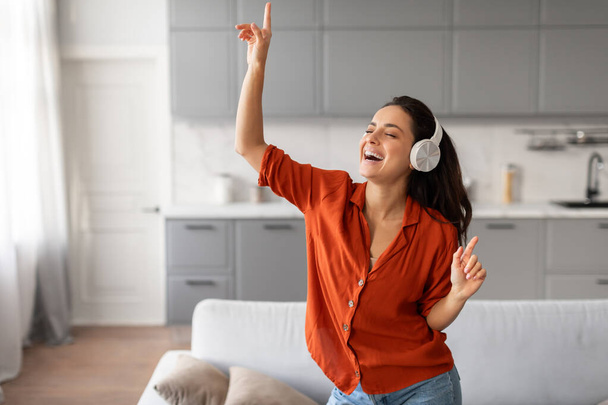 Femme insouciante et heureuse en chemise orange vibrante danse dans son salon, profitant de ses airs préférés avec des écouteurs élégants, plein de vie, espace libre - Photo, image
