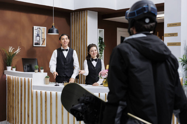 At front desk of exclusive winter resort, two receptionists warmly welcoming male guest in snow gear. Tourist wearing winter jacket and carrying snowboarding equipment arrives in hotel lobby. - Photo, Image