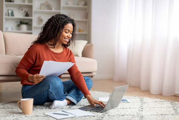 Sorrindo preto mulher freelancer trabalhando com laptop e documentos em casa, feliz jovem afro-americano fêmea sentada no chão no quarto, verificando documentos e usando o computador, desfrutando de trabalho remoto - Foto, Imagem