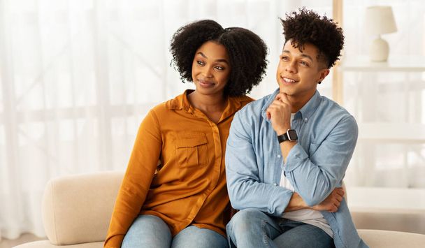 Frohes afroamerikanisches Millennial-Paar sitzt eng auf dem Sofa, Frau und Mann lächeln nachdenklich, beide genießen einen entspannten und gemütlichen Moment zusammen in einem lichtdurchfluteten Raum - Foto, Bild