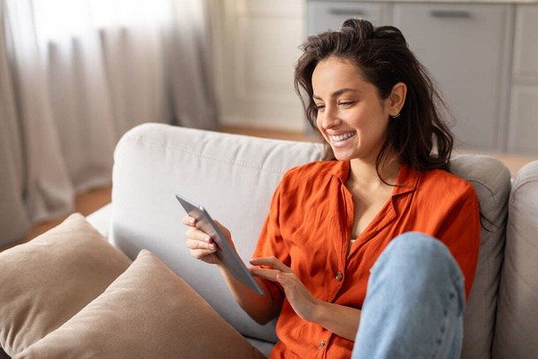 Cheerful young woman comfortably lounging on her couch, deeply engaged with her tablet, enjoying relaxed day in the comfort of her stylish home - Photo, Image