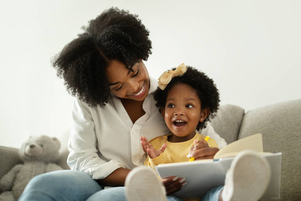 Мила маленька афроамериканська дівчинка з луком для волосся насолоджується розвиваючими заняттями зі своєю мамою, плескаючи руками і сміючись. Щаслива чорна мати і дочка малюють разом, домашній інтер'єр - Фото, зображення