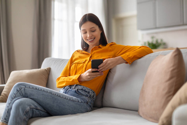 Улыбающаяся молодая женщина удобно сидит на диване, погруженная в свой смартфон, одетая в яркую желтую рубашку и джинсы, сидя в уютной, естественно освещенной гостиной - Фото, изображение