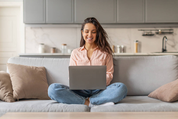 Szczęśliwa młoda kobieta freelancer wygodnie siedzi na szarej sofie, pracując na laptopie z uśmiechem, w jasnym, współczesnym salonie, ilustrując zdalną pracę - Zdjęcie, obraz