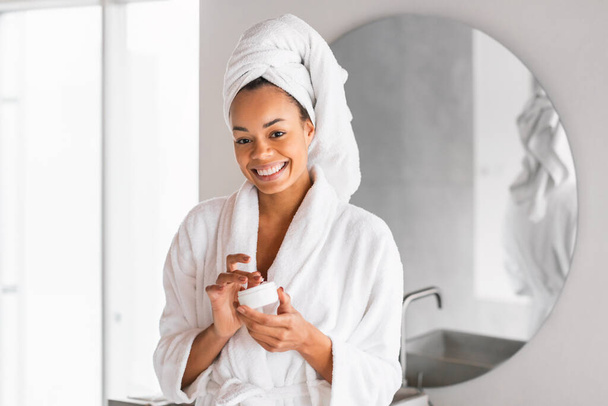 Szczęśliwa czarna kobieta ze zdrową i świecącą cerą z ulubionym kremem nawilżającym, reklamowym produktem urody, stojąca w szlafroku z ręcznikiem na głowie w nowoczesnej łazience - Zdjęcie, obraz