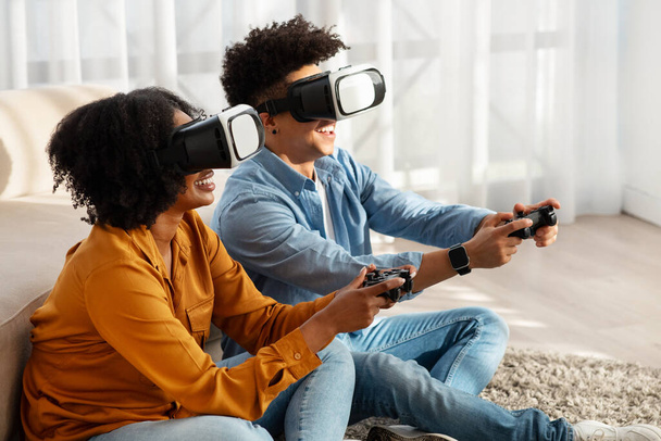 Ενθουσιασμένο ζευγάρι Αφροαμερικανών της χιλιετίας βυθισμένο σε ένα παιχνίδι εικονικής πραγματικότητας, φορώντας ακουστικά VR και χειριστήρια συγκράτησης, καθισμένο σε ένα μαλακό χαλί σε ένα ηλιόλουστο σαλόνι στο σπίτι - Φωτογραφία, εικόνα
