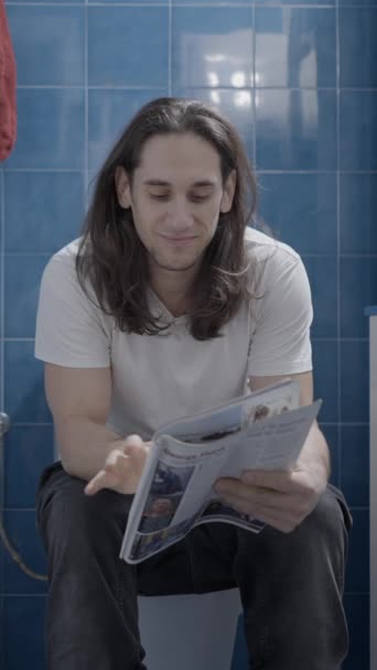 Young caucasian man reading a newsletter in the bathroom while shit - Frontal shot - Vertical video - Πλάνα, βίντεο