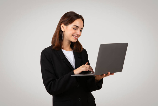 Empresária caucasiana engajada em um blazer preto com uma camisa branca sorrindo enquanto ela digita em um laptop, exibindo produtividade e conectividade em um ambiente profissional - Foto, Imagem