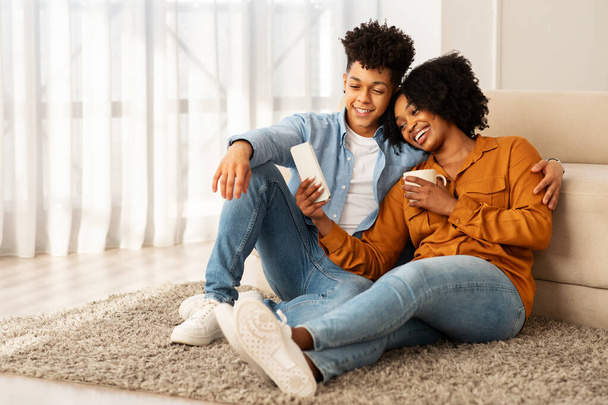 Ένα ζεστό νεαρό ζευγάρι αφροαμερικανών με casual ένδυμα, χαλαρώνει άνετα σε ένα βελούδινο χαλί, μοιράζεται μια οθόνη smartphone και απολαμβάνει μια ζεστή, οικεία στιγμή στο φωτεινό σαλόνι τους - Φωτογραφία, εικόνα