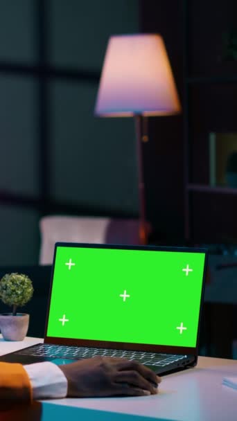 Вертикальное видео Африканская молодая женщина смотрит на ноутбук с зеленым экраном, сидит за своим домашним столом и готовится к посещению онлайн университетских уроков. Девушка, использующая компьютер с изолированным макетом - Кадры, видео