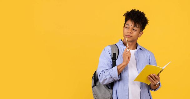 Studente maschio nero contemplativo con lo zaino che tiene la matita al mento e al notebook, perso nel pensiero sullo sfondo giallo, incarnando attenzione e curiosità accademica - Foto, immagini