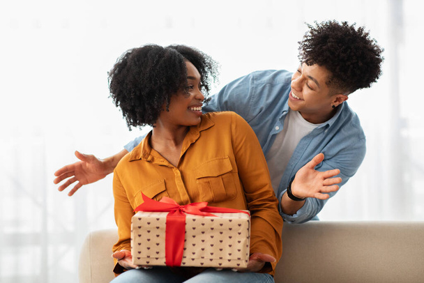 Podekscytowana afro-amerykańska milenijna kobieta otrzymuje prezent z wzorami serca od uśmiechniętego mężczyzny, tworząc radosny i intymny moment dawania i niespodzianki w dobrze oświetlonym pokoju - Zdjęcie, obraz