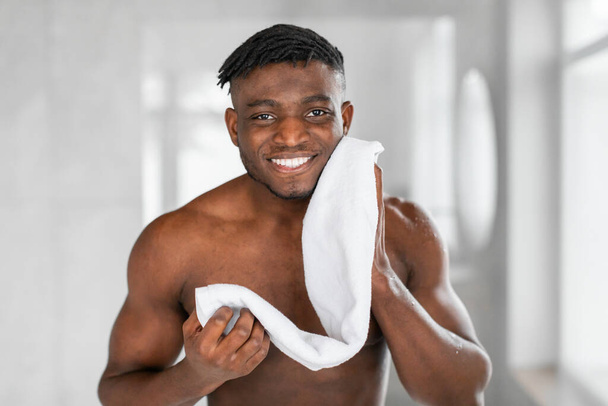Porträt eines fröhlichen schwarzen Millennial-Mannes, der sein Gesicht im modernen Badezimmer mit einem Handtuch wischt, ohne Hemd dasteht und in die Kamera lächelt. Männliche Hautpflege und männliches Pflegekonzept - Foto, Bild