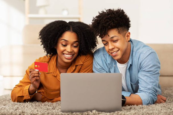 Feliz pareja afroamericana tumbada en el suelo con un ordenador portátil, mujer con una tarjeta de crédito, ambos participando alegremente en compras en línea en su cómoda y elegante sala de estar - Foto, Imagen