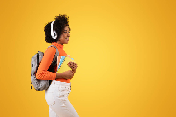 Πλευρική άποψη ενός νεαρού Αφροαμερικανού μαθητή με σγουρά μαλλιά που φορούσε άσπρα ακουστικά, πορτοκαλί ζιβάγκο και γκρι σακίδιο, κρατώντας σημειωματάρια σε κίτρινο φόντο - Φωτογραφία, εικόνα