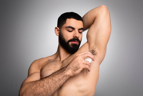 Uomo del Medio Oriente che applica deodorante antitraspirante su ascella in piedi su sfondo grigio. Igiene maschile routine, sudore e odore di protezione, cosmetici per la cura della pelle - Foto, immagini