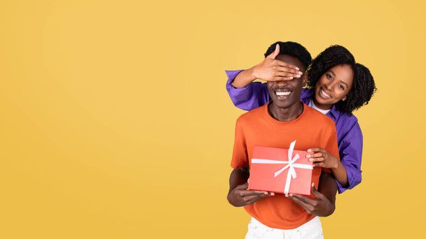 A smiling woman in a purple shirt playfully covers the eyes of a man in an orange shirt, who is holding a red gift box with a white ribbon, both against a bright yellow background - Photo, Image