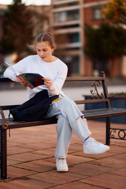 Dans l'ambiance sereine du parc, une belle adolescente blonde se plonge dans un livre, trouvant réconfort et stimulation mentale en attendant le début de sa journée scolaire..  - Photo, image