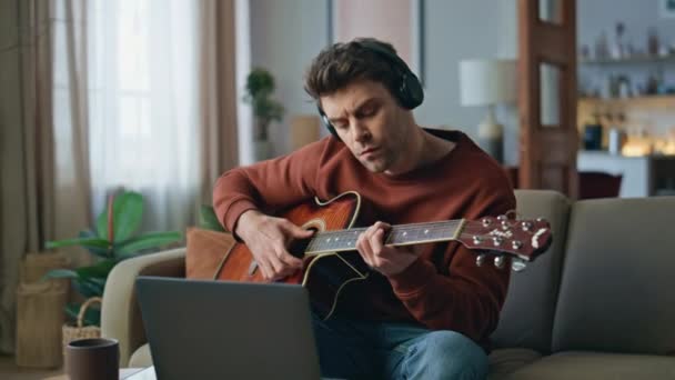 自宅のソファーでノートパソコンのウェブカメラでオンラインレッスンを録音する笑顔のギタリスト. イヤホンの音を聞くギターを弾く才能のあるハンサムな男. 楽器音楽を練習する幸せな男性ミュージシャン. - 映像、動画