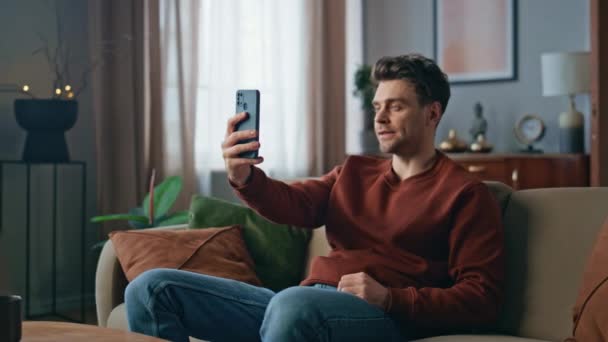 Handsome guy recording self video on smartphone at modern living room. Confident attractive man looking cellphone web camera at online call. Relaxed hipster creating content for social media on phone - Footage, Video