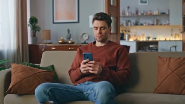自宅のソファーで携帯電話のアプリケーションを閲覧する深刻な男. 現代のアパートでソファーに座っているスマートフォン画面にタイピングするハンサムな若者. 携帯電話アプリを使用してオンライン注文を重視した顧客. - 映像、動画