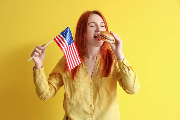Joven pelirroja con bandera de EE.UU. comiendo hamburguesa sobre fondo amarillo - Foto, Imagen