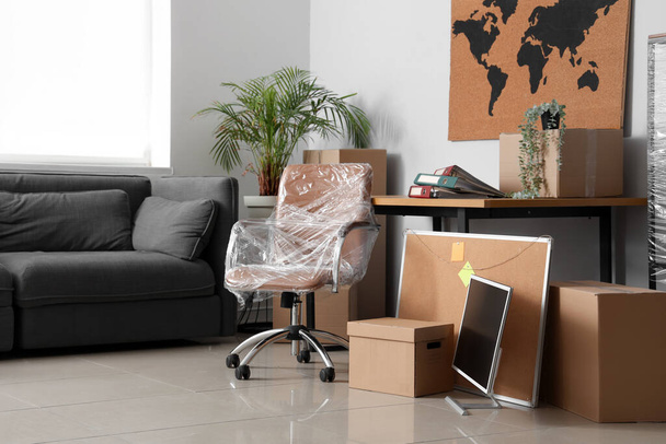 Intérieur du bureau avec chaise enveloppée dans un film étirable, canapé et boîtes en carton le jour du déménagement - Photo, image
