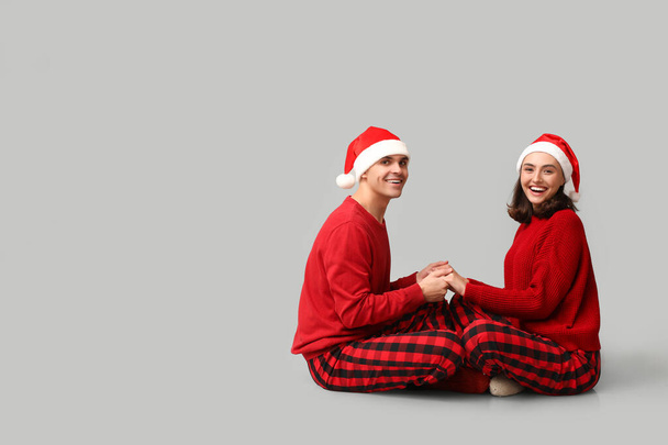 Joyeux jeune couple en pyjama de Noël et chapeaux de Père Noël assis sur fond gris - Photo, image
