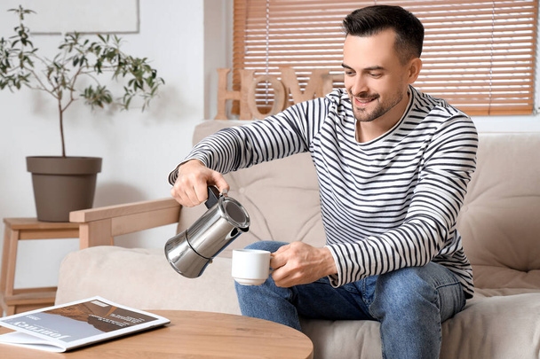 Knappe man schenkt espresso van geiser koffiezetapparaat in beker in woonkamer - Foto, afbeelding