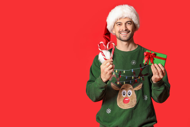 Hombre guapo en sombrero de Santa con regalo de Navidad y bastones de caramelo sobre fondo rojo - Foto, Imagen