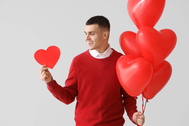 Όμορφος άντρας με χάρτινη καρδιά και μπαλόνια σε ανοιχτό φόντο. Γιορτή του Αγίου Βαλεντίνου - Φωτογραφία, εικόνα