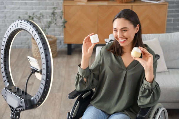 Vrouwelijke blogger in rolstoel met potten crème opname video thuis - Foto, afbeelding