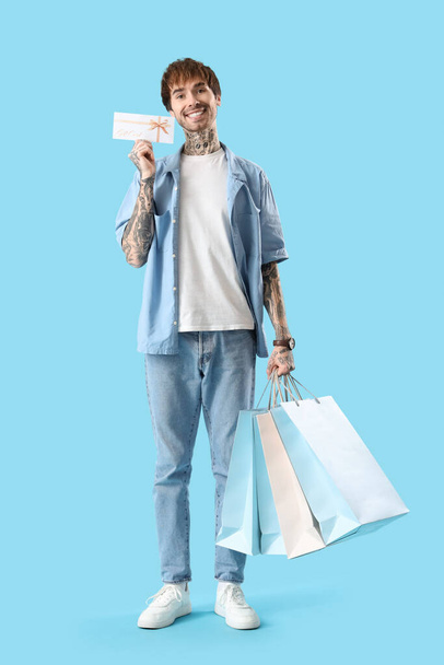 Молодий татуйований чоловік з подарунковою карткою та сумками на синьому фоні - Фото, зображення