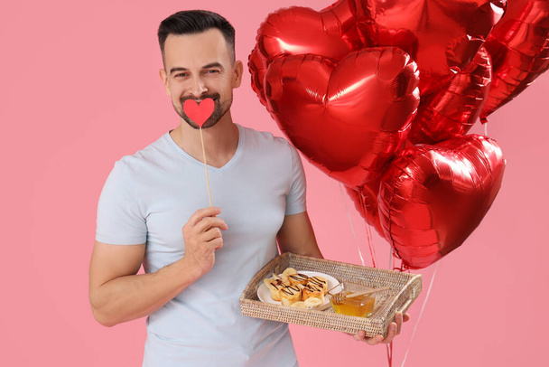 Young man with tasty breakfast and heart-shaped balloons on pink background. Valentine's Day celebration - Photo, Image