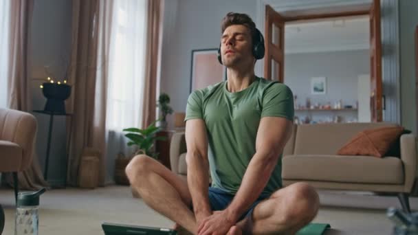 Peaceful athlete practicing meditation at home yoga mat in wireless headphones. Tranquil man closing eyes listening tutorial audio at tablet. Serene guy making breathing exercises in earphones indoors - Footage, Video