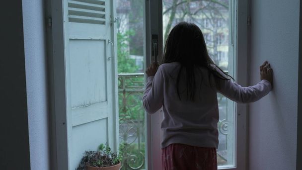 Achterzijde van een duur kind dat bij het raam staat en vanuit het appartement op de tweede verdieping naar het uitzicht staart in humeurig melancholisch. Bedachtzaam klein meisje leunend op glas - Foto, afbeelding